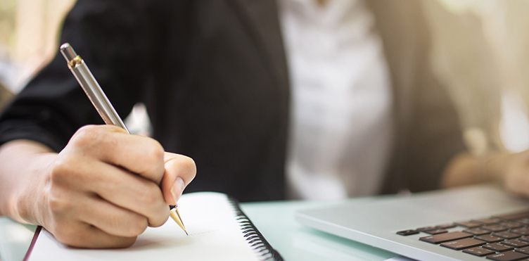 Photo of an individual at a desk with a laptop and writing in a notebook